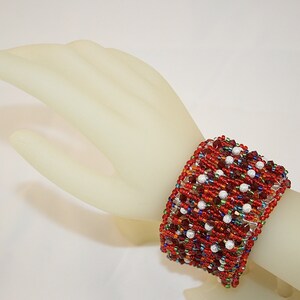 May include: A red, white, and multi-colored beaded bracelet with a woven design. The bracelet is on a mannequin's wrist.