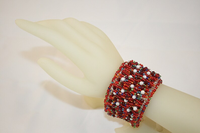 May include: A red, white, and multi-colored beaded bracelet with a woven design. The bracelet is on a mannequin's wrist.