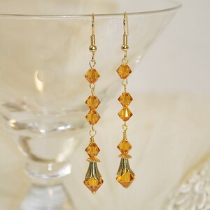 May include: A pair of gold dangle earrings with amber-colored crystal beads. The earrings have a teardrop shape with a gold-toned metal accent at the bottom.