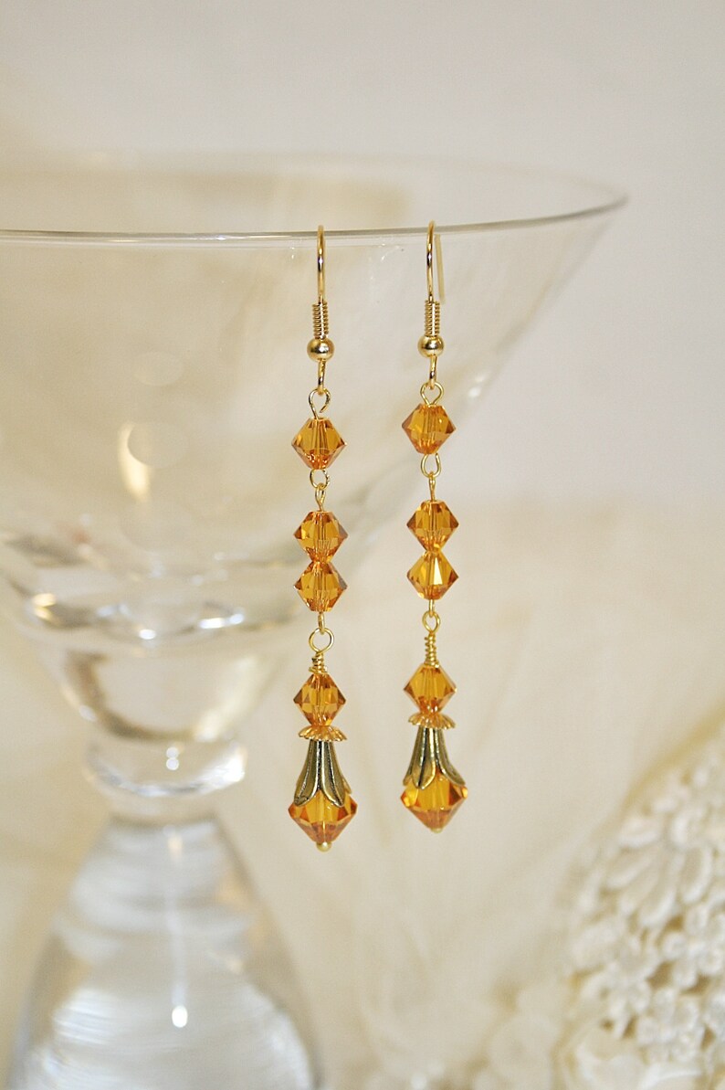 May include: A pair of gold dangle earrings with amber-colored crystal beads. The earrings have a teardrop shape with a gold-toned metal accent at the bottom.