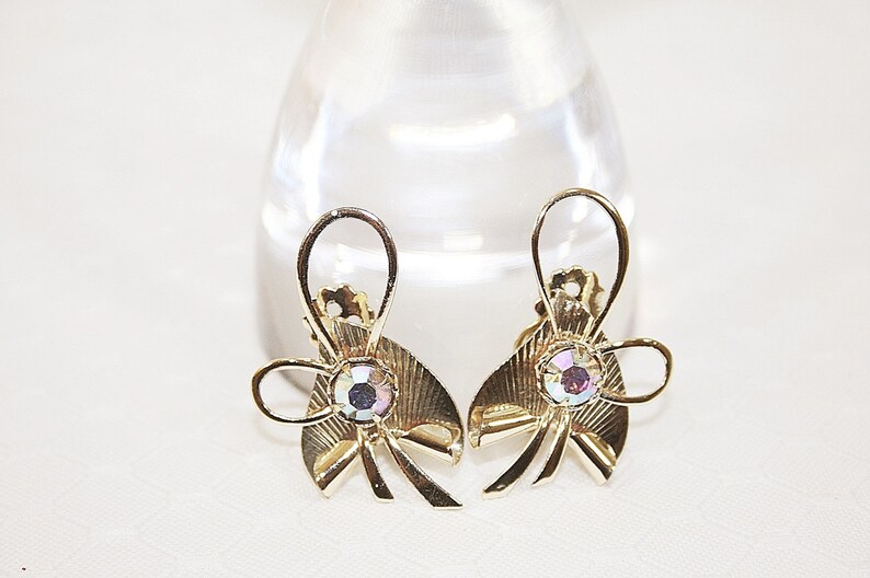 May include: A pair of gold tone bow-shaped earrings with iridescent rhinestones in the center.