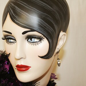 May include: A mannequin head with a vintage hairstyle, dramatic eye makeup, and red lipstick. The mannequin wears a silver earring with a small, decorative charm. A black and purple feather boa is around the neck.
