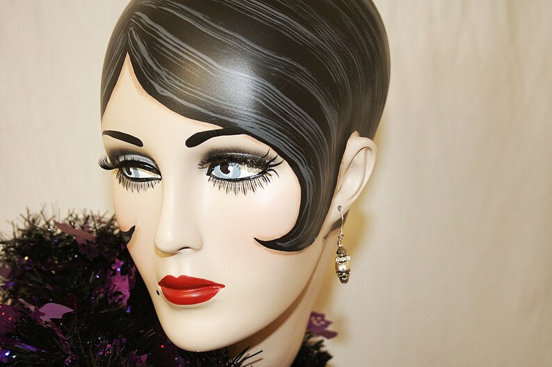 May include: A mannequin head with a vintage hairstyle, dramatic eye makeup, and red lipstick. The mannequin wears a silver earring with a small, decorative charm. A black and purple feather boa is around the neck.