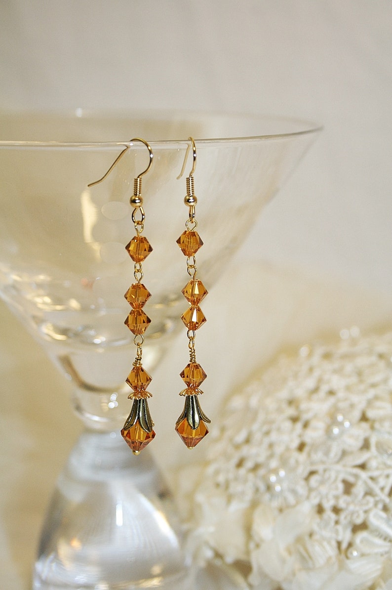 May include: A pair of long, gold dangle earrings with amber-colored crystal beads. The earrings have a teardrop-shaped crystal at the bottom.