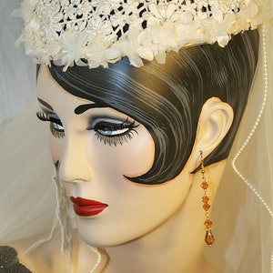 May include: A white crocheted hat with a floral design, worn by a mannequin with dark hair and a veil. The mannequin is wearing a pearl necklace and amber earrings.
