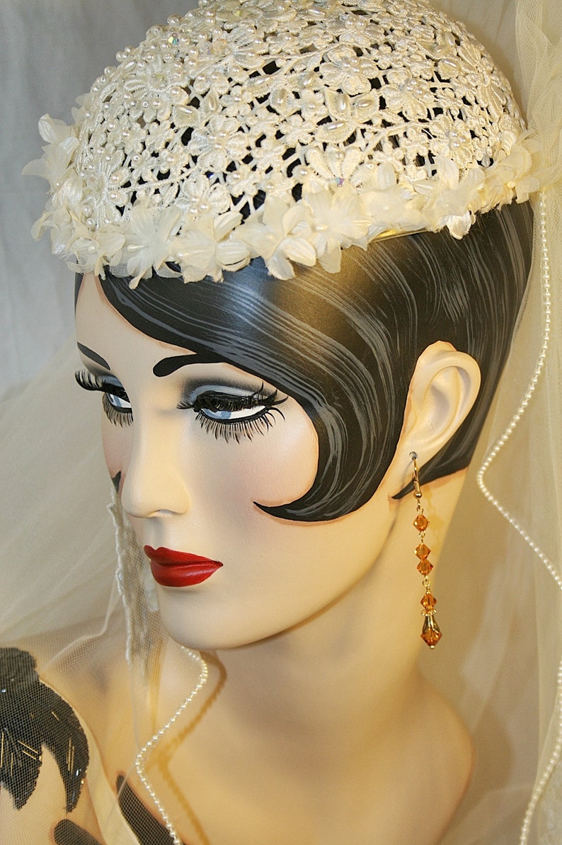 May include: A white crocheted hat with a floral design, worn by a mannequin with dark hair and a veil. The mannequin is wearing a pearl necklace and amber earrings.