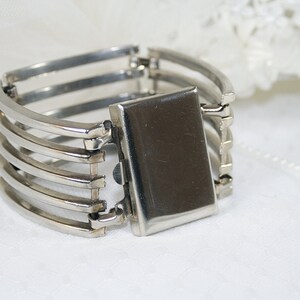 May include: A silver bracelet with a rectangular, flat, silver clasp. The bracelet is made of multiple bands of silver that are connected together.