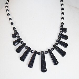 Puede incluir: Un collar blanco y negro con un dise&ntilde;o en forma de abanico de colgantes de piedras preciosas negras. El collar est&aacute; hecho con peque&ntilde;as cuentas blancas y cuentas de piedras preciosas negras.