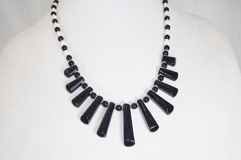 Puede incluir: Un collar blanco y negro con un dise&ntilde;o en forma de abanico de colgantes de piedras preciosas negras. El collar est&aacute; hecho con peque&ntilde;as cuentas blancas y cuentas de piedras preciosas negras.
