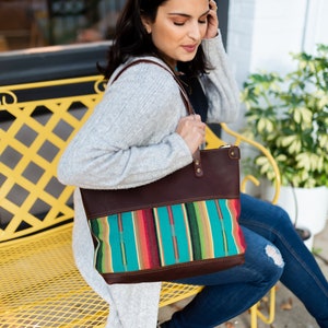 May include: A woman sits on a yellow bench holding a brown leather tote bag with a colorful woven fabric panel. The bag has a zipper closure and a top handle.