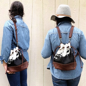 May include: Two people are wearing a black and white cowhide bucket bag. One person is wearing the bag as a backpack and the other is wearing it as a shoulder bag. Both bags have brown leather trim.