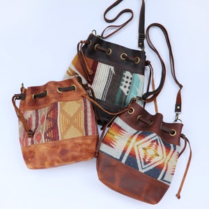 May include: Three bucket bags featuring brown leather bases and tops with drawstring closures. Each bag displays a unique woven textile panel with geometric patterns in shades of orange, red, blue, cream, and brown, complemented by adjustable leather straps.