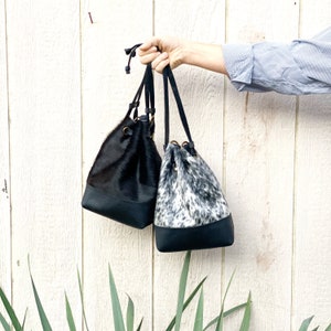 May include: Two black leather bucket bags with drawstrings. One bag is black with a black and white cowhide pattern. The other bag is black with a solid black cowhide pattern.