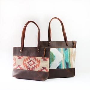 May include: Two brown leather tote bags with patterned fabric panels. The bags have brown leather handles and bases. One bag features a geometric design in red, orange, and beige. The other has a turquoise, green, and beige pattern.