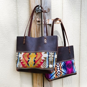 May include: Two leather tote bags with colorful woven fabric panels. The larger bag has a geometric pattern in shades of red, yellow, blue, and brown. The smaller bag has a geometric pattern in shades of blue, purple, and white.