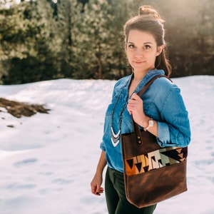 May include: A brown leather tote bag featuring a horizontal band with a geometric pattern in shades of orange, cream, green, and black. The bag has two leather shoulder straps attached with brass-colored metal rivets and a rectangular body shape.