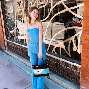 May include: A light blue denim jumpsuit with thin straps and a square neckline, featuring a fitted bodice and wide-leg trousers with front pockets. The jumpsuit is paired with a black handbag featuring a geometric pattern in turquoise, white, and brown.