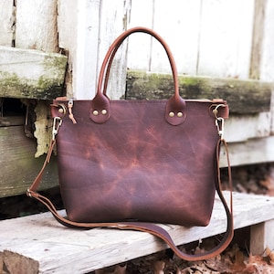 May include: A brown leather tote bag with a long strap and brass hardware. The bag has a distressed look and is sitting on a wooden surface.