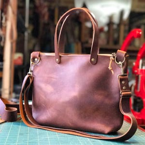 May include: A brown leather tote bag with a zip closure and a long adjustable strap. The bag has a worn, vintage look.