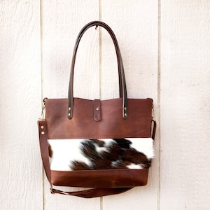 May include: A brown leather tote bag with a white and brown cowhide patch. The bag has two top handles and a long shoulder strap.