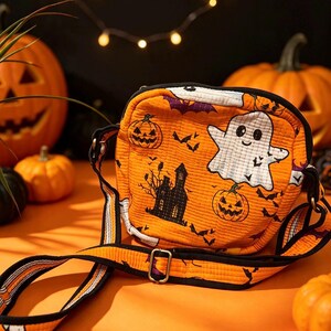 May include: A quilted orange crossbody bag featuring a Halloween-themed print with white ghosts, jack-o'-lanterns, black haunted houses, and bats. The bag has a black trim, a zip closure, and an adjustable strap with a metal buckle.