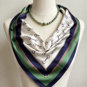 May include: A square silk scarf featuring a white centre with a repeating black horse bit pattern, bordered by wide navy blue and green stripes with brown leather-like strap details. A necklace of round green beads rests above the scarf.