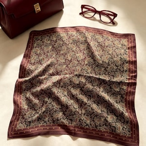 May include: A square scarf featuring a dark brown and cream paisley pattern with a solid dark red border, accompanied by a matching dark red structured handbag with a gold clasp and a pair of dark red framed eyeglasses.