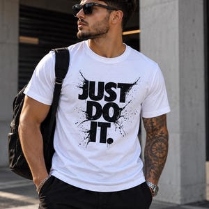 Può includere: Una t-shirt bianca a maniche corte con la scritta nera JUST DO IT in uno stile grafico a schizzi di vernice usurata sul petto. Il capo presenta un girocollo ed è abbinato a uno zaino nero e a pantaloni cargo neri.