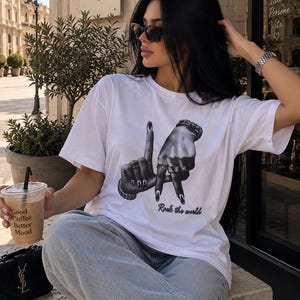 Può includere: Una t-shirt bianca a maniche corte con una stampa grafica in bianco e nero di due mani che formano le lettere L e A, con il testo "Rock the world" stampato sotto in un carattere corsivo. La maglietta ha un girocollo.