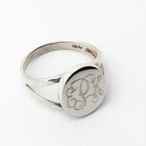 May include: A silver-toned ring featuring a circular face engraved with the cursive monogram letters EPK. The band has a split design that connects to the sides of the circular face. The interior of the band shows a small hallmark stamp.