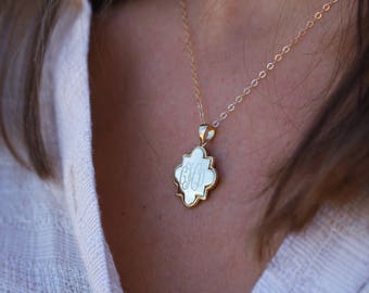 Puede incluir: Un collar de cadena de oro con un colgante de oro con un monograma. El colgante tiene forma de estrella de cuatro puntas con una superficie lisa y blanca.