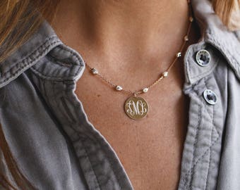 Puede incluir: Un collar dorado con un colgante redondo con iniciales grabadas. El collar tiene una cadena fina con pequeños detalles de cuentas. El colgante cuelga sobre el pecho de una persona que lleva una camisa gris abotonada.
