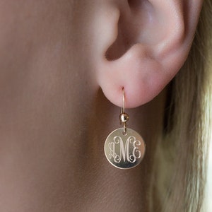 May include: Gold dangle earrings with a round disc charm featuring a monogram of the letters "M.C.E."