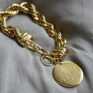 May include: A gold chain bracelet with a large, round, gold charm. The charm has a monogram engraved on it.