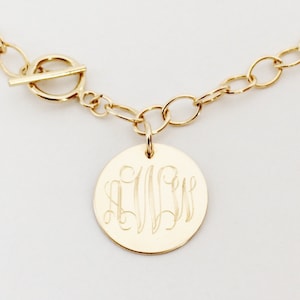 May include: A gold chain necklace with a round pendant featuring a monogram. The chain has a toggle clasp.