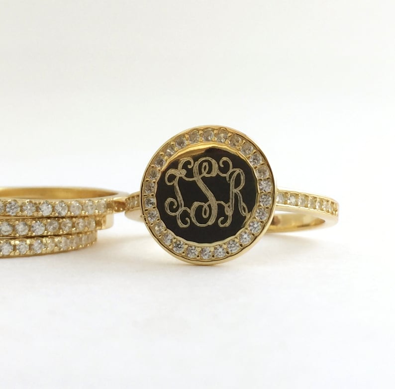 Puede incluir: Un anillo de oro con una cara circular que tiene una incrustaci&oacute;n negra grabada con las letras LSR en letra cursiva. La cara circular est&aacute; bordeada por peque&ntilde;as piedras claras y la banda tambi&eacute;n tiene una hilera de piedras claras en la parte superior.