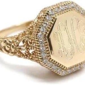 May include: A gold ring featuring an octagonal face with an engraved monogram, surrounded by a halo of small clear stones. The band displays intricate filigree metalwork along the sides, extending from the central setting to the shank.