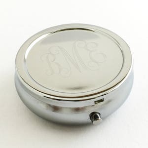 May include: A silver round pill box with a hinged lid. The lid has a monogrammed inscription of the letters "B.V.E."