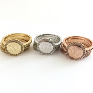 Puede incluir: Tres anillos apilados en tonos oro, plata y oro rosa. Cada anillo presenta una cara circular con un monograma grabado en letra cursiva, rodeado por un halo de peque&ntilde;as piedras claras. Las bandas tambi&eacute;n est&aacute;n incrustadas con piedras.