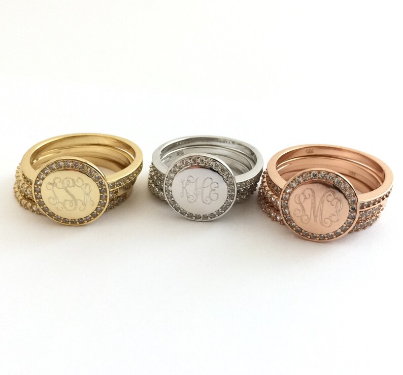 Puede incluir: Tres anillos apilados en tonos oro, plata y oro rosa. Cada anillo presenta una cara circular con un monograma grabado en letra cursiva, rodeado por un halo de peque&ntilde;as piedras claras. Las bandas tambi&eacute;n est&aacute;n incrustadas con piedras.