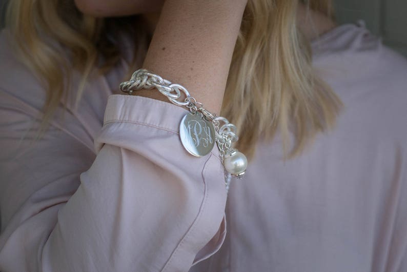 Puede incluir: Un brazalete de plata con un gran dije redondo grabado con las iniciales "PM" y un dije de perla. El brazalete se lleva en la mu&ntilde;eca.
