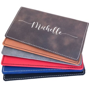 May include: A stack of six personalized faux leather checkbook covers in various colors, including gray, brown, red, blue, and black. The top cover is personalized with the name "Michelle" in a white script font.