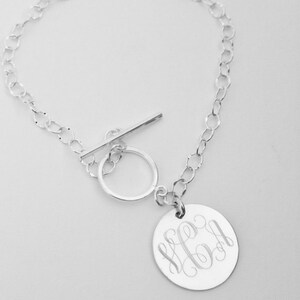 May include: A silver-colored bracelet with a toggle clasp and a circular pendant. The pendant is engraved with a monogram in a cursive font. The bracelet chain is made of small, interconnected links.