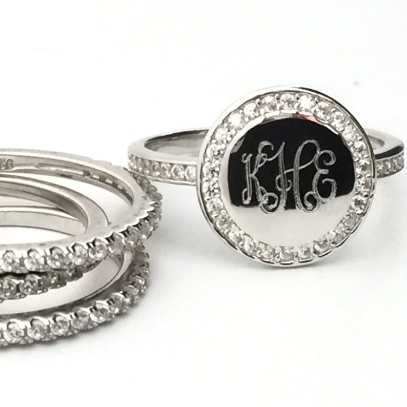 Puede incluir: Un conjunto de anillos de tono plateado con una pieza central circular grabada con las letras KHE. La pieza central est&aacute; rodeada por un halo de peque&ntilde;as piedras claras, y las bandas tambi&eacute;n est&aacute;n incrustadas con piedras claras en sus bordes.