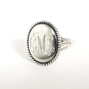 May include: A silver-toned ring featuring a large, oval-shaped face with a polished, smooth surface. The center of the oval is engraved with the stylized, interconnected letters CMH. A decorative, twisted rope-style border frames the oval face.