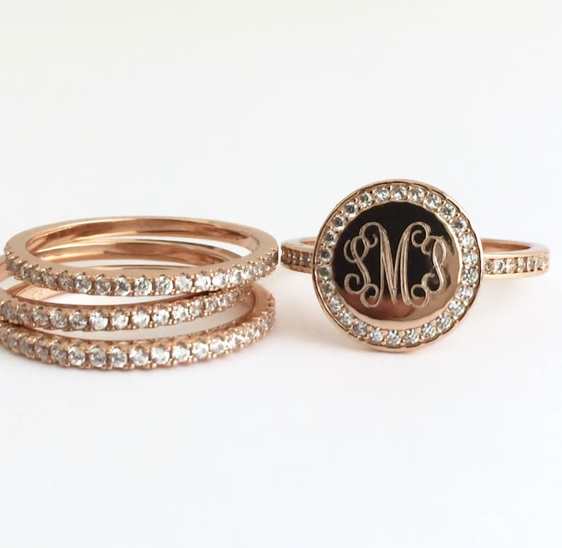 Puede incluir: Un conjunto de anillos de oro rosa con una pieza central circular grabada con las letras SMS en letra cursiva. La cara circular y las bandas de los anillos est&aacute;n incrustadas con peque&ntilde;as piedras redondas y transparentes que reflejan la luz.