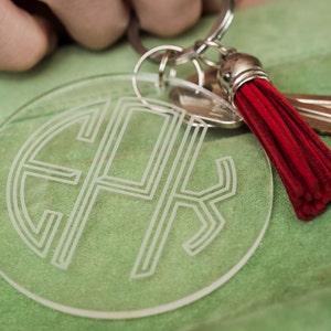 May include: A clear acrylic keychain with a monogrammed design and a red tassel. The keychain is attached to a silver key ring.