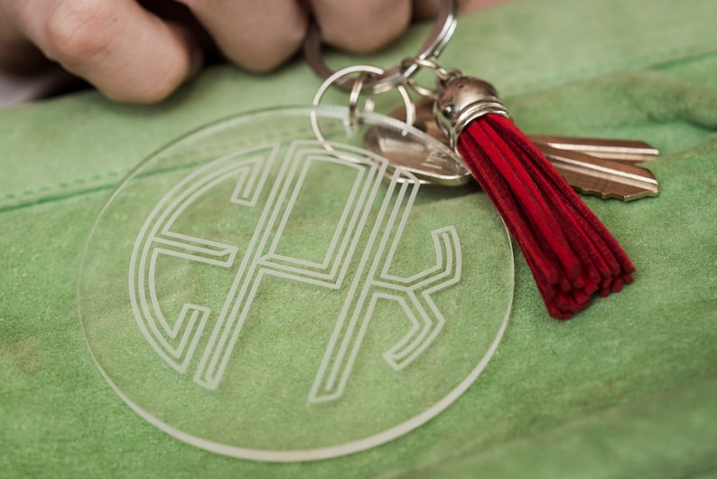 May include: A clear acrylic keychain with a monogrammed design and a red tassel. The keychain is attached to a silver key ring.