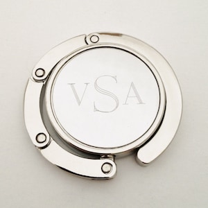 May include: Silver-toned metal bag hook with a circular design. The hook is engraved with the initials "VSA".