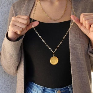 May include: A gold chain necklace with a large, round pendant featuring a monogram. The pendant is engraved with the letters "GBS". The necklace is worn by a person wearing a brown cardigan and a black tank top.